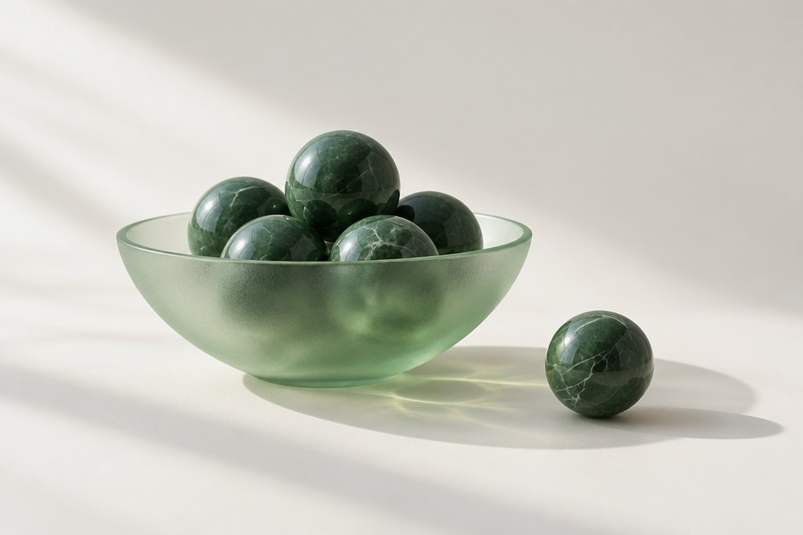 A wide shallow frosted glass bowl filled with green marble spheres, with one more sphere resting beside it, on a bright studio surface